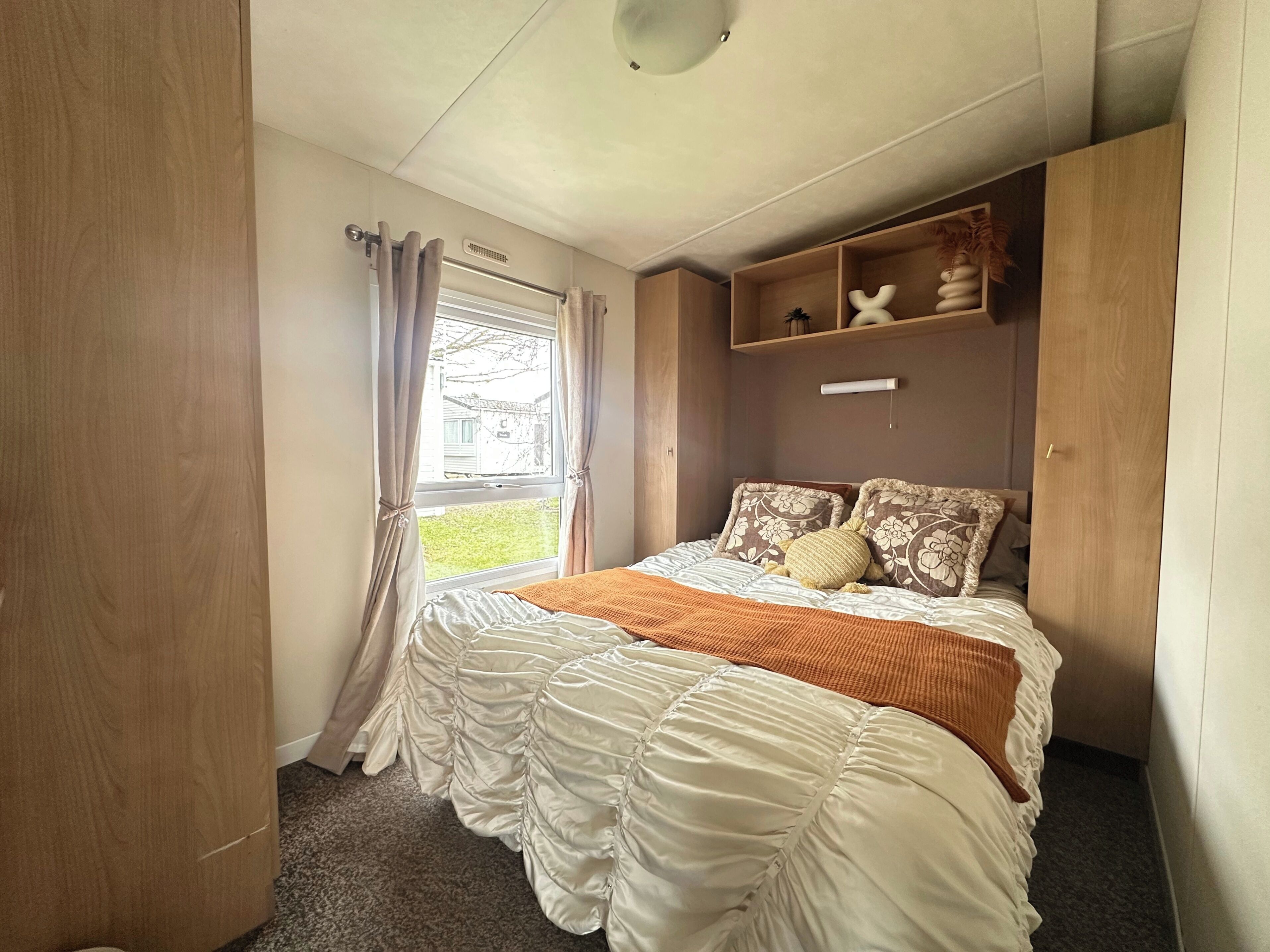 A bedroom with a large bed, a window with curtains, a shelf with decorative items, and a door.