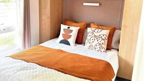 A neatly made bed with a quilted white and orange blanket, two pillows with abstract designs, and a wooden shelf above the bed with decorative items. - Harwich - Caravan - Sleeps 10 - Garden - Pool (Harwich)