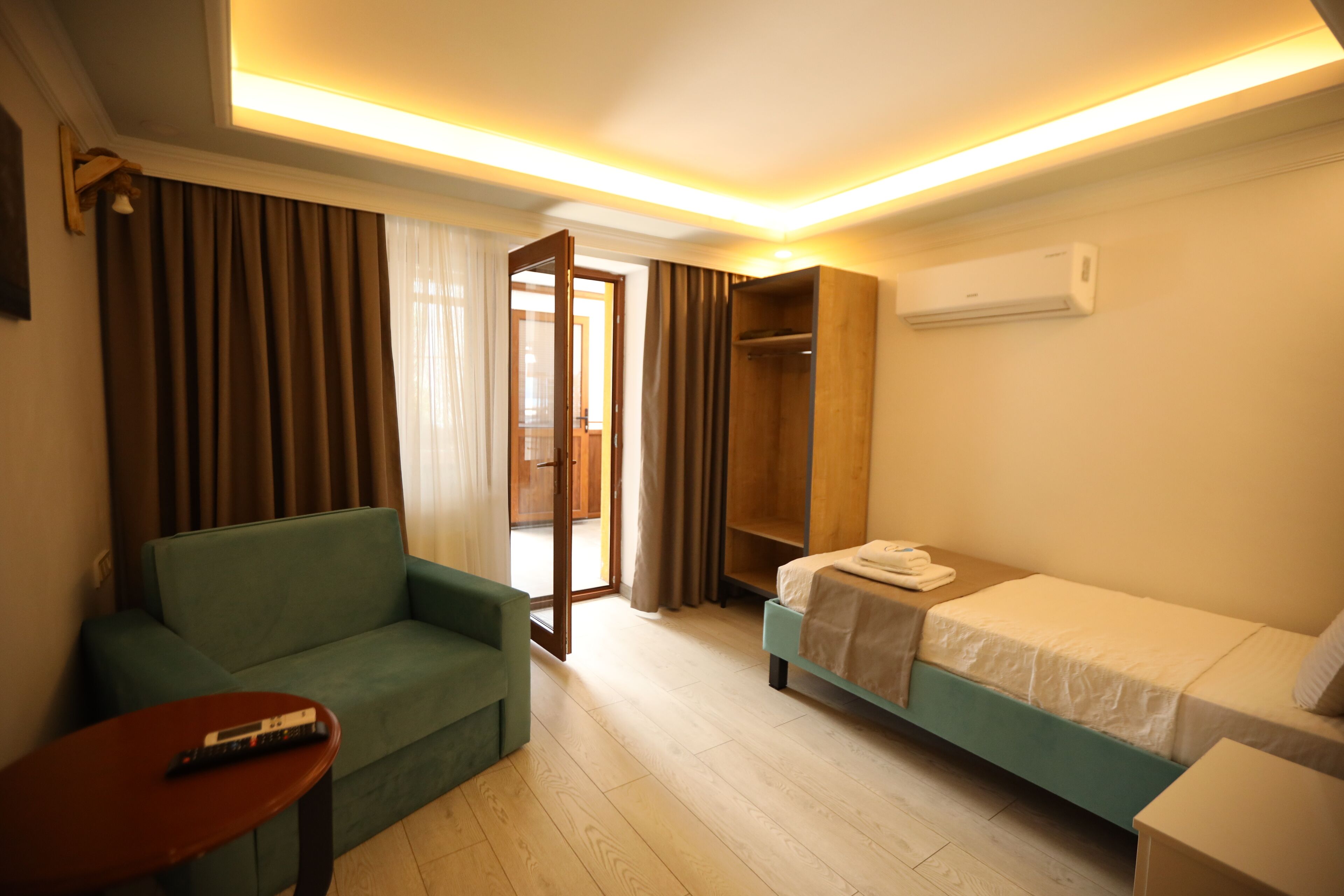 A hotel room with a sofa, a single bed, a small table, a wardrobe, and an air conditioning unit.
