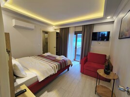 A hotel room with a bed, a red armchair, a small table with a plant, a TV mounted on the wall, and a sliding glass door leading to an outdoor area.