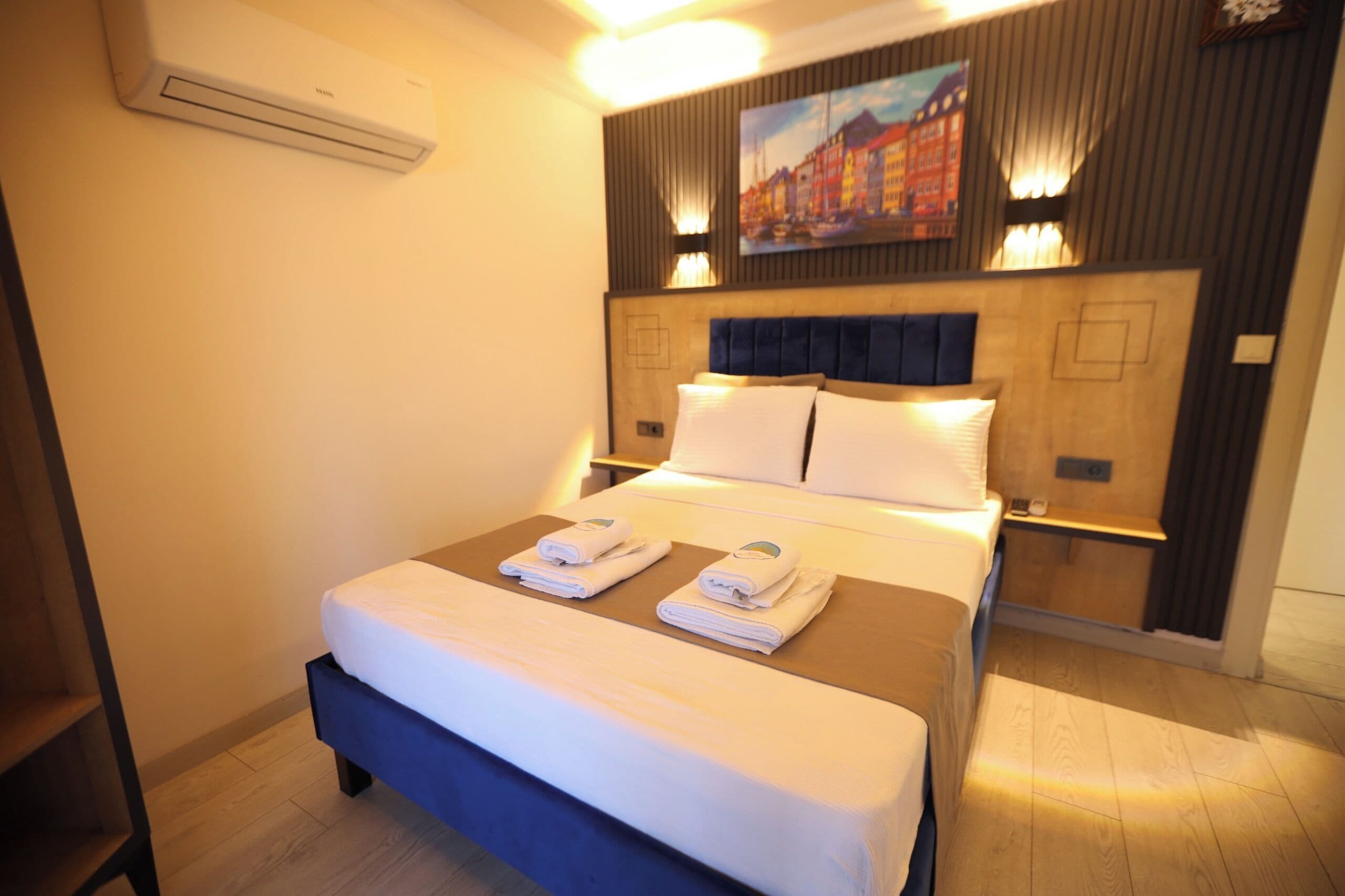 A hotel room with a bed, bedside tables, a wall-mounted air conditioner, and a painting above the bed.