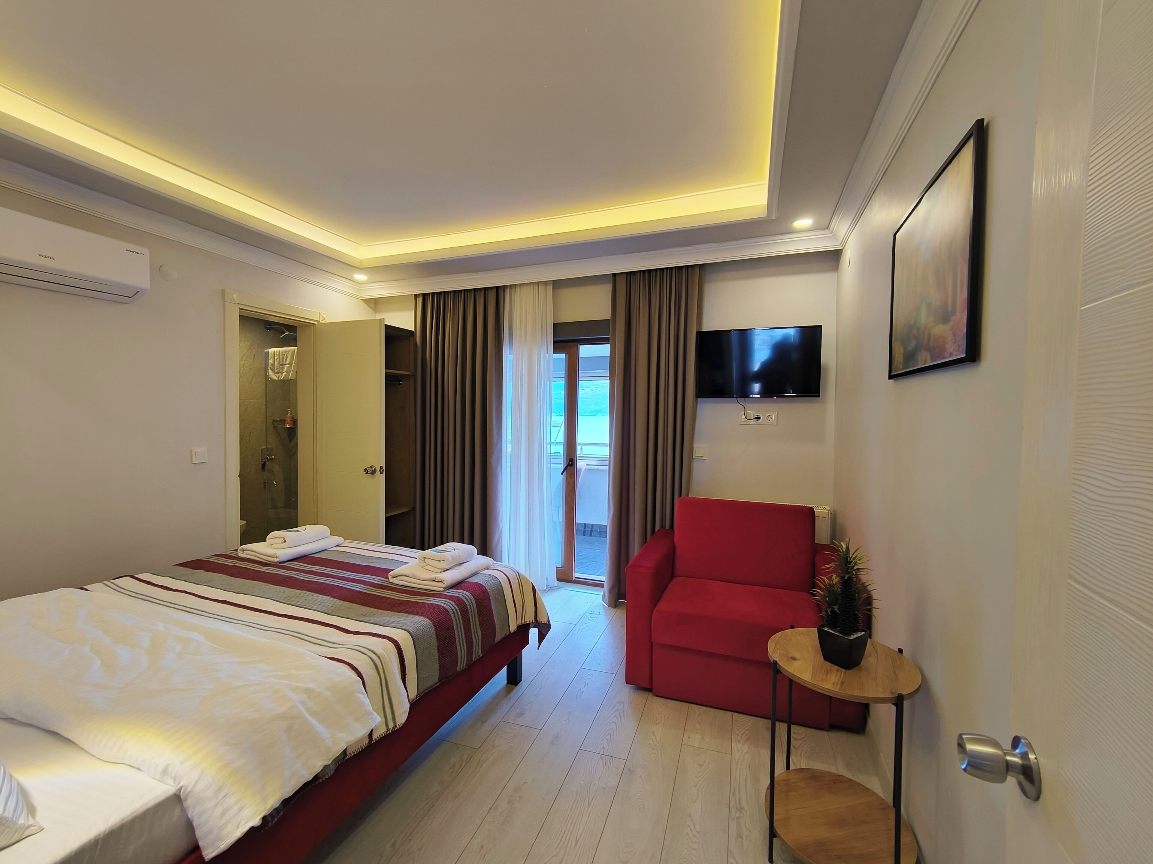 A hotel room with a large bed, a red armchair, a small table with a plant, a TV mounted on the wall, and a door leading to a balcony.