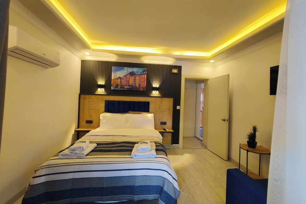 A modern hotel room with a large bed, a wooden headboard, a painting above the bed, and a small table with a plant.