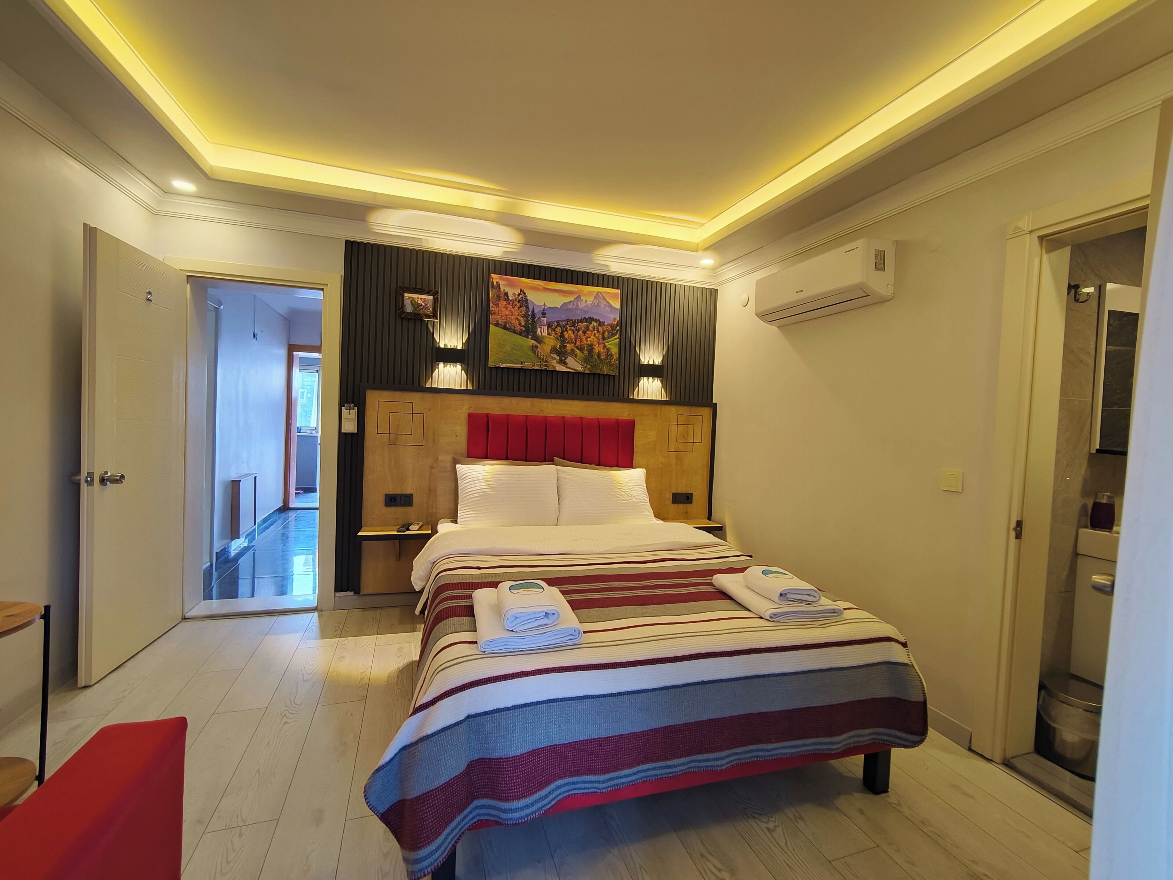 Habitación de hotel con cama, mesita de noche, un banco rojo, suelo de madera y un pasillo que lleva a otra habitación.