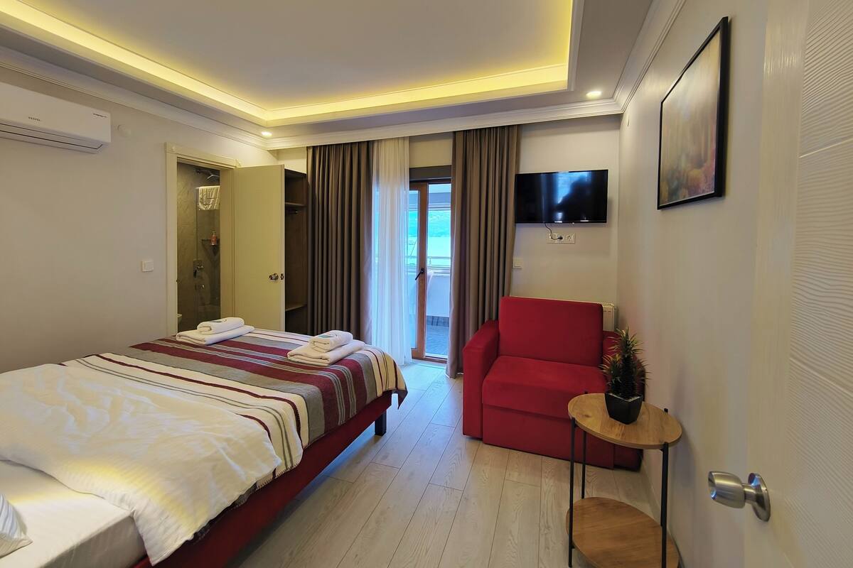 A hotel room with a bed, a red chair, a TV, and a balcony door.