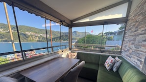 A room with a view of a lake and hills, a wooden table, two chairs, and a green sofa with floral cushions. - AMASRA EMEK PANSİYON (Amasra)