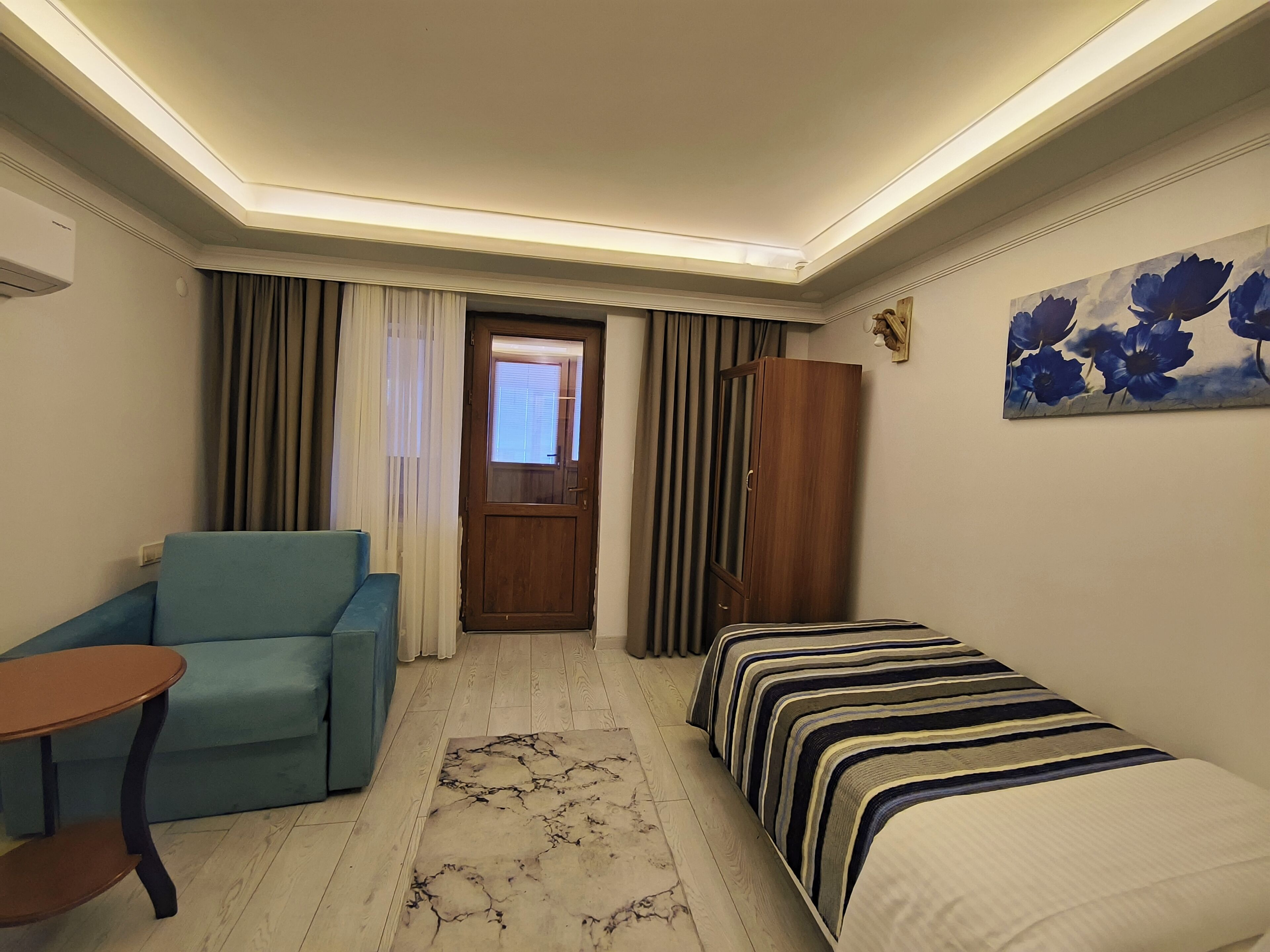 A compact hotel room with a single bed, a sofa, a small table, a wooden wardrobe, and a window with curtains.