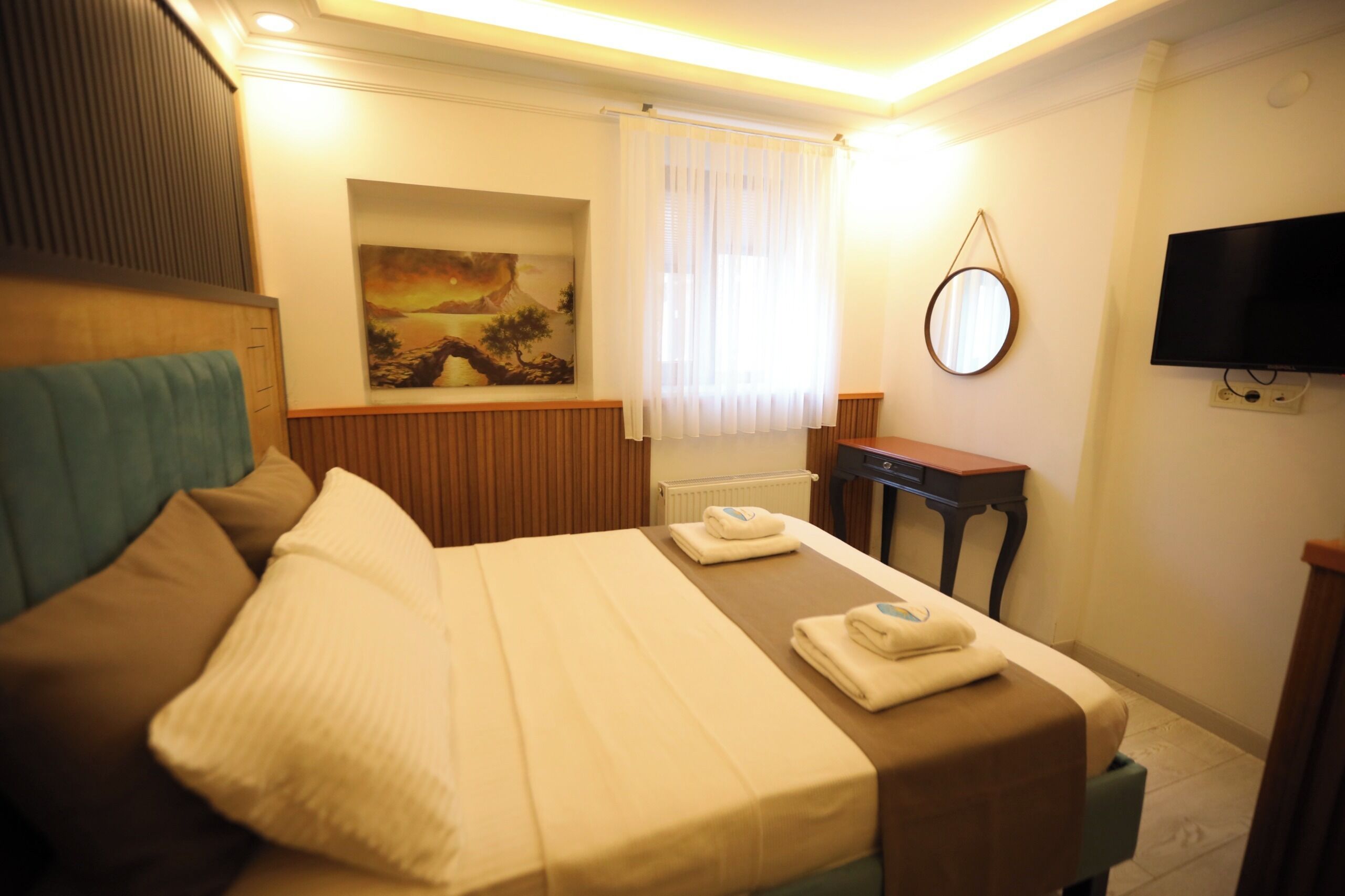 Una habitación de hotel con cama, escritorio, televisor, un cuadro y ventana con cortinas.