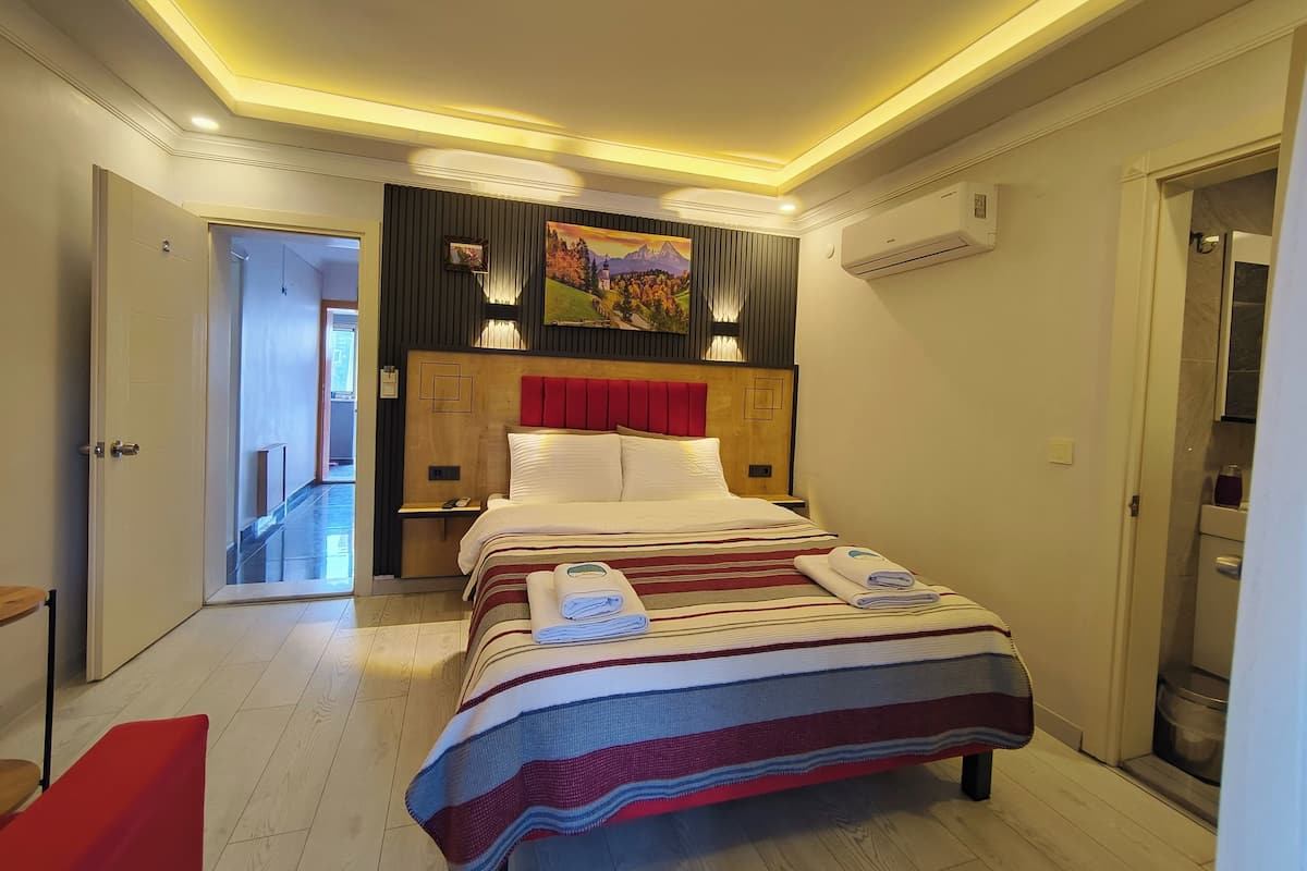 A hotel room with a bed, a nightstand, a red bench, a wooden floor, and a hallway leading to another room.
