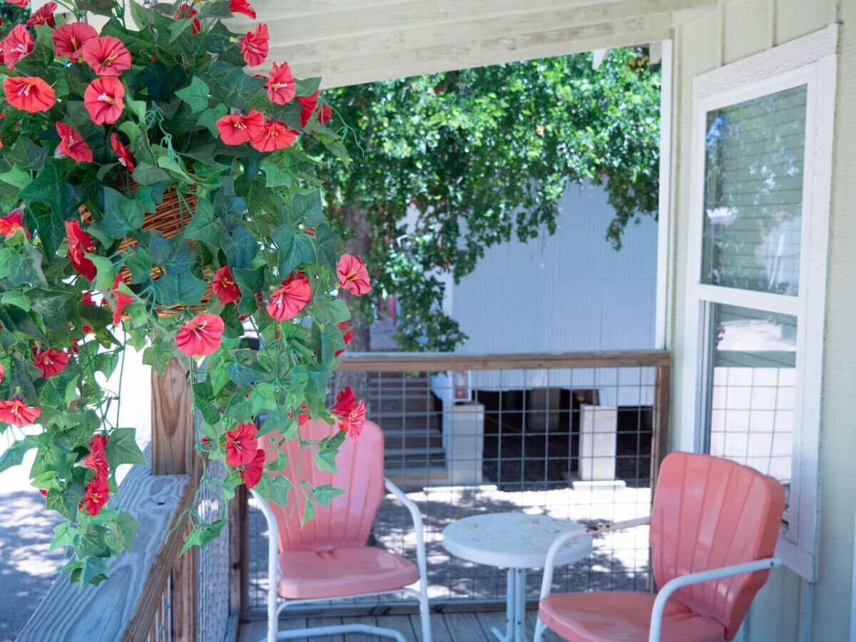 A covered patio with two pink chairs and a table, surrounded by a wooden fence and a hanging plant with red flowers.