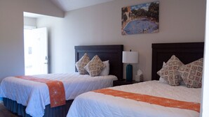 A hotel room with two beds, each with a dark headboard, white bedding, and patterned pillows. There is a painting on the wall above the beds, and a door leading to another room. - River View Double Queen Cottage near Downtown #24 (Kerrville)