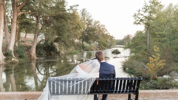 A couple sitting on a bench by a river, surrounded by trees.