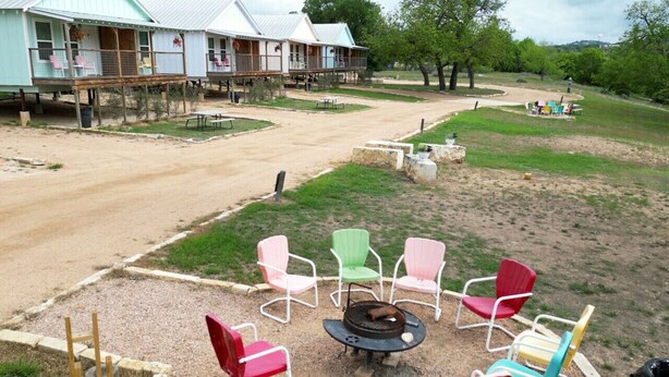 A rustic lodge with a dirt path leading to a seating area with colorful chairs and a fire pit. - Single Level Double Queen Beds Cabin 2 w/ Carport (Kerrville)