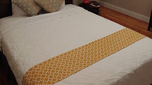 Una cama bien tendida con una colcha blanca y anaranjada estampada, dos almohadas con motivos y una lámpara de noche roja, además de un aire acondicionado de pared.