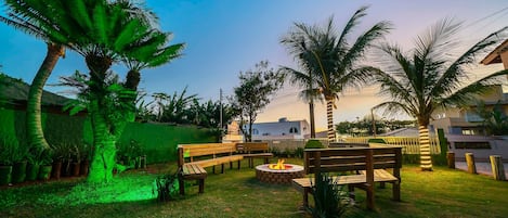 Espaço ao ar livre com bancos de madeira e lareira, cercado por palmeiras e uma cerca verde.