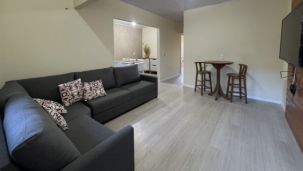 Una sala de estar moderna con un sofá seccional, una zona de comedor con dos sillas y una mesa pequeña, y un televisor de pantalla plana montado en la pared.