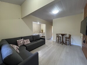 Una sala de estar moderna con un sofá seccional, una zona de comedor con dos sillas y una mesa pequeña, y un televisor de pantalla plana montado en la pared.