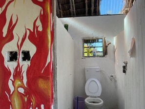 A bathroom with a white toilet, a small window, and a wall with a red and yellow abstract design. - Charming beach-front cottage in Tiwi for easy-going friends with AC and WiFi (Tiwi)