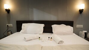 A neatly made bed with white linens, two wall-mounted light fixtures, a bedside table with a kettle and a fan, and a power outlet. - Maha Kumbh Mandapam  (Karchana)