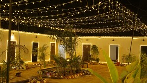 A courtyard with string lights, palm trees, and a building with doors. - Maha Kumbh Mandapam  (Karchana)