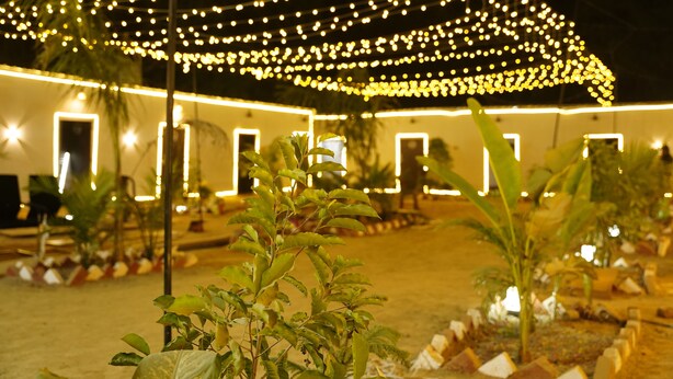 An outdoor area with string lights, potted plants, and a building with illuminated outlines. - Maha Kumbh Mandapam  (Karchana)