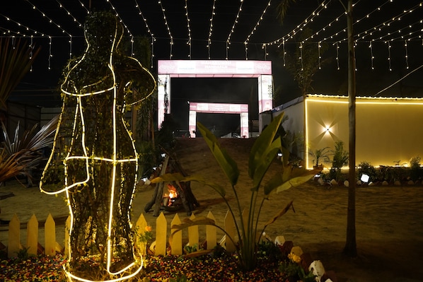 A nighttime outdoor area with a decorative light sculpture, a fire pit, and a large illuminated archway. - Maha Kumbh Mandapam  (Karchana)