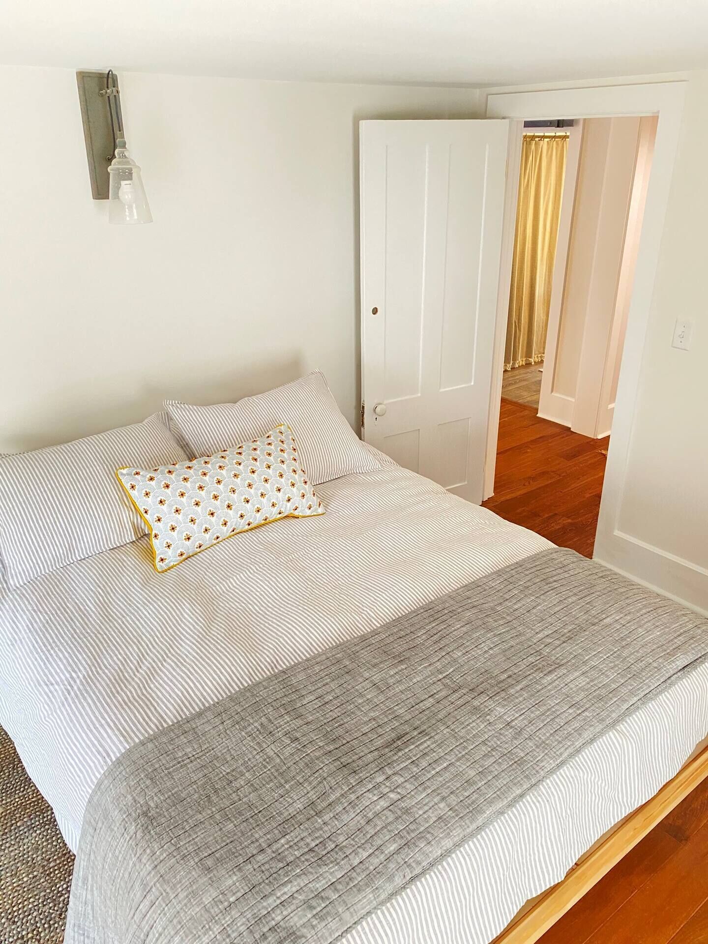 Una cama bien tendida con ropa de cama a rayas y una almohada estampada, un aplique en la pared y una puerta que da a otra habitación.