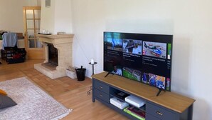 A living room with a flat-screen TV mounted on the wall, a fireplace, a wooden cabinet below the TV, and a patterned rug on the floor. - Lovely 3-star country house, ideal for a family. (LEUTENHEIM)