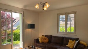 A living room with a brown leather sofa, a wooden coffee table, a floor lamp, and a large window with a view of greenery and a house. - Lovely 3-star country house, ideal for a family. (LEUTENHEIM)
