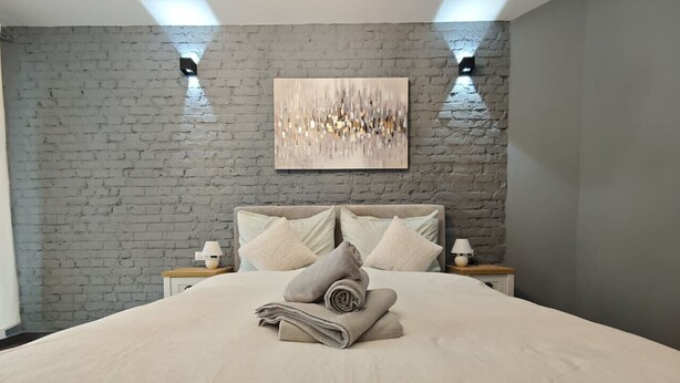 Una cama bien hecha con una colcha color beige, dos mesitas de noche con lámparas, una pared de ladrillo y un cuadro abstracto moderno sobre la cama.