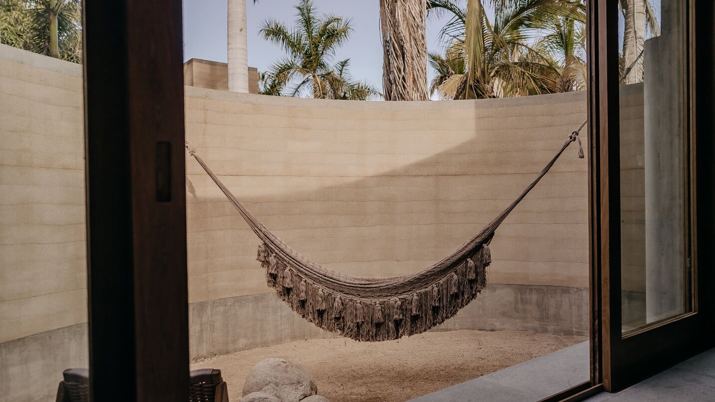 Una hamaca colgada entre dos paredes en una habitación con suelo de arena, palmeras al fondo y una puerta de cristal.