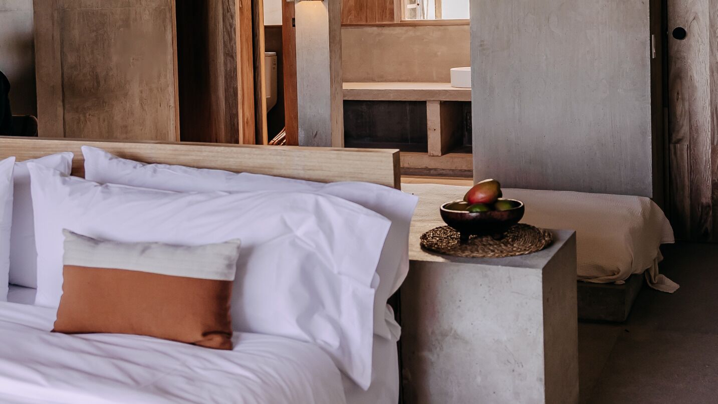 Un lit avec du linge de lit blanc et un oreiller marron, une petite table en bois sur laquelle se trouve un bol de fruits, et un pilier en béton.