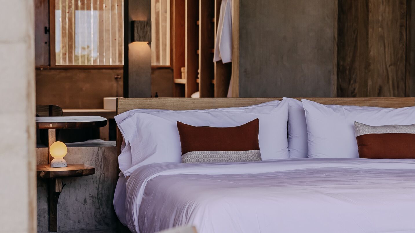Un lit avec du linge de lit blanc et des oreillers marron, une petite table de chevet avec une lampe, et une vue sur un mur et une fenêtre en bois.