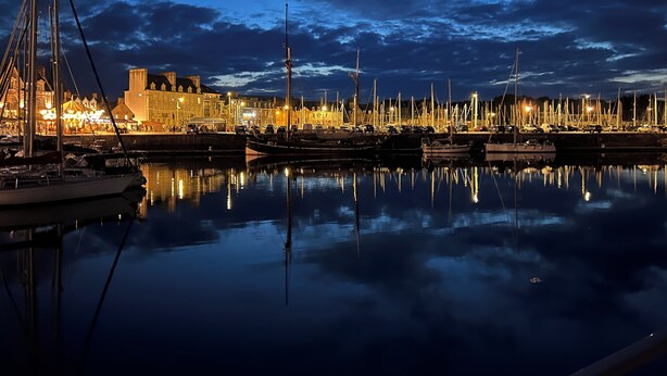 Un port de plaisance au crépuscule, avec des bateaux amarrés, une eau calme qui reflète le paysage et des bâtiments en arrière-plan.