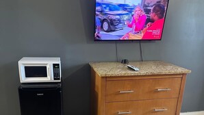 Een kamer met een aan de muur bevestigde televisie, een kleine zwarte koelkast, een houten dressoir en een bed met een kleurrijke deken.