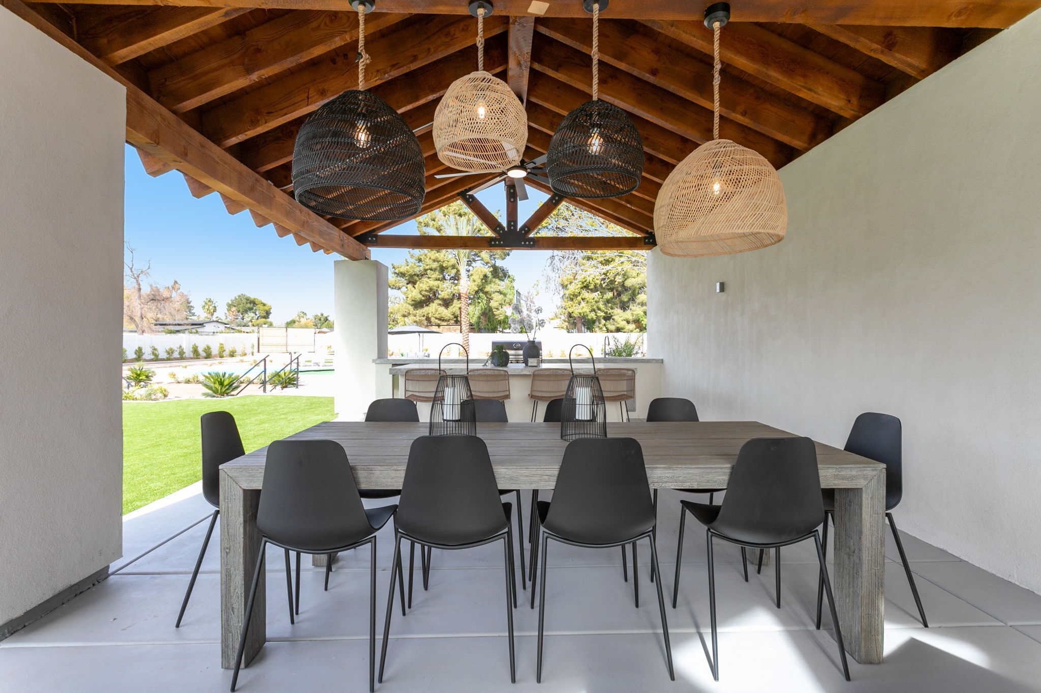 A covered outdoor dining area with a wooden table and black chairs, a ceiling fan, and hanging wicker lamps.