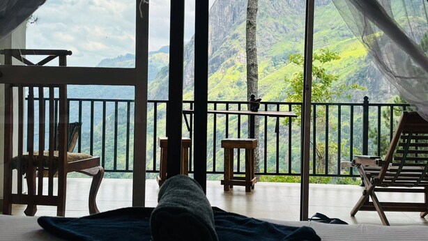 Una stanza con una grande finestra che si affaccia su un paesaggio montuoso. Si vede un letto con una coperta scura, una sedia di legno e un tavolino con una statua.