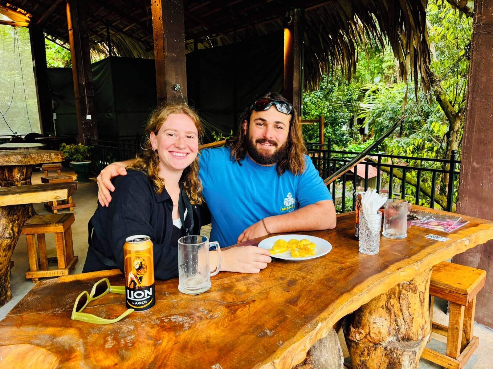 Two people seated at a wooden table with a beer can, a glass, and a plate of food.