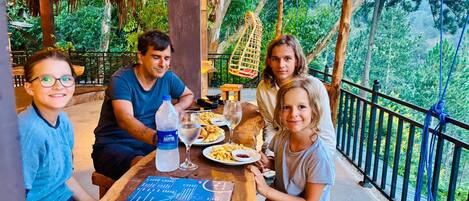 A family dining at an outdoor table with a menu, surrounded by lush greenery and a thatched roof structure.