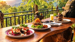 Un tavolo di legno con piatti di cibo, un bicchiere di succo e uno zaino su un balcone con vista su vegetazione e montagne.