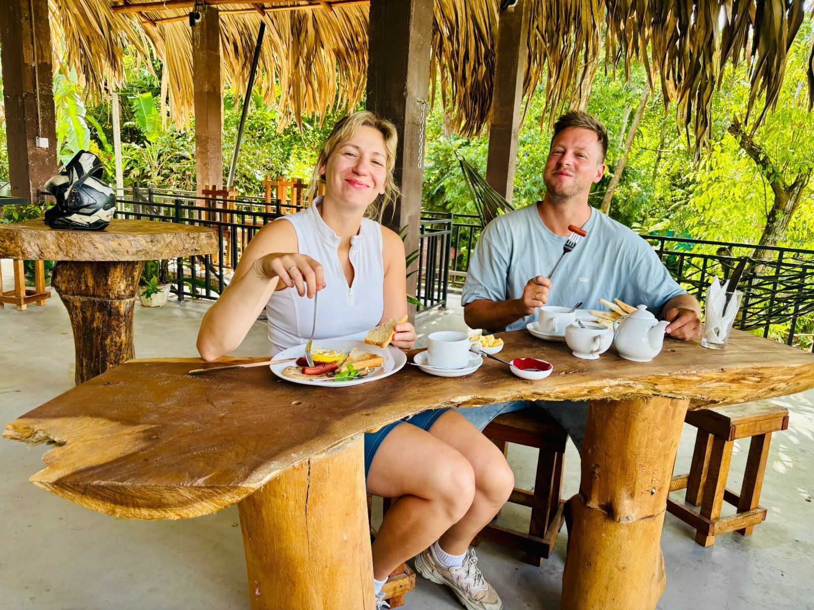 Two people dining at a wooden table with a thatched roof overhead, surrounded by lush greenery.