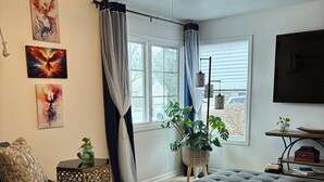 Ett vardagsrum med en takfläkt, ett stort fönster med gardiner, en tv som är monterad på väggen och en krukväxt.
