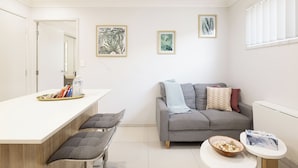 A modern living room with a grey sofa, a white coffee table, and a small round table with a bowl and tissue box. There is a white counter with a tray of snacks and a water bottle. - Smart Suites Self Check-in Apartments - 39 Freney (Rocklea)