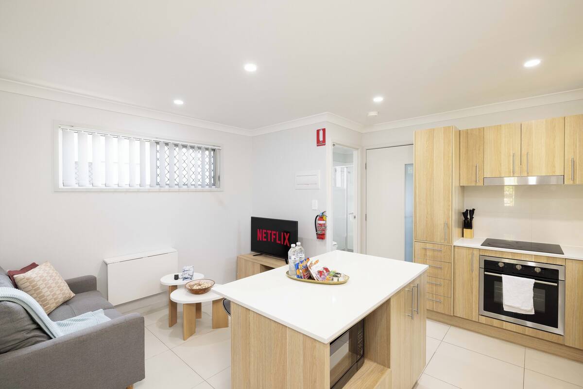 A modern kitchen with a central island, wooden cabinets, and a built-in oven. There is a small dining area with a table and chairs, and a sofa in the living area.