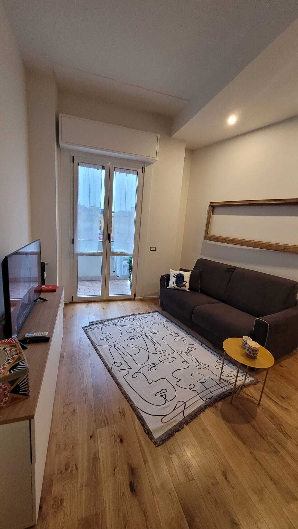 Un saló amb terra de fusta, un sofà marró, una taula rodona petita, un televisor sobre un suport i una finestra gran amb vista a l'exterior.