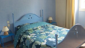 Une chambre avec un lit bleu, deux lampes fixées au mur, une fenêtre avec des rideaux et deux tableaux encadrés accrochés au mur.