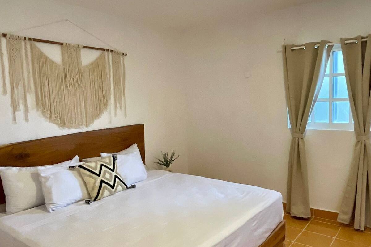 A neatly made bed with white linens and a wooden headboard, a small potted plant on the nightstand, and a window with beige curtains.