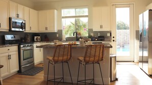 Una cocina moderna con armarios blancos, electrodomésticos de acero inoxidable, un ventanal amplio y dos taburetes de bar.