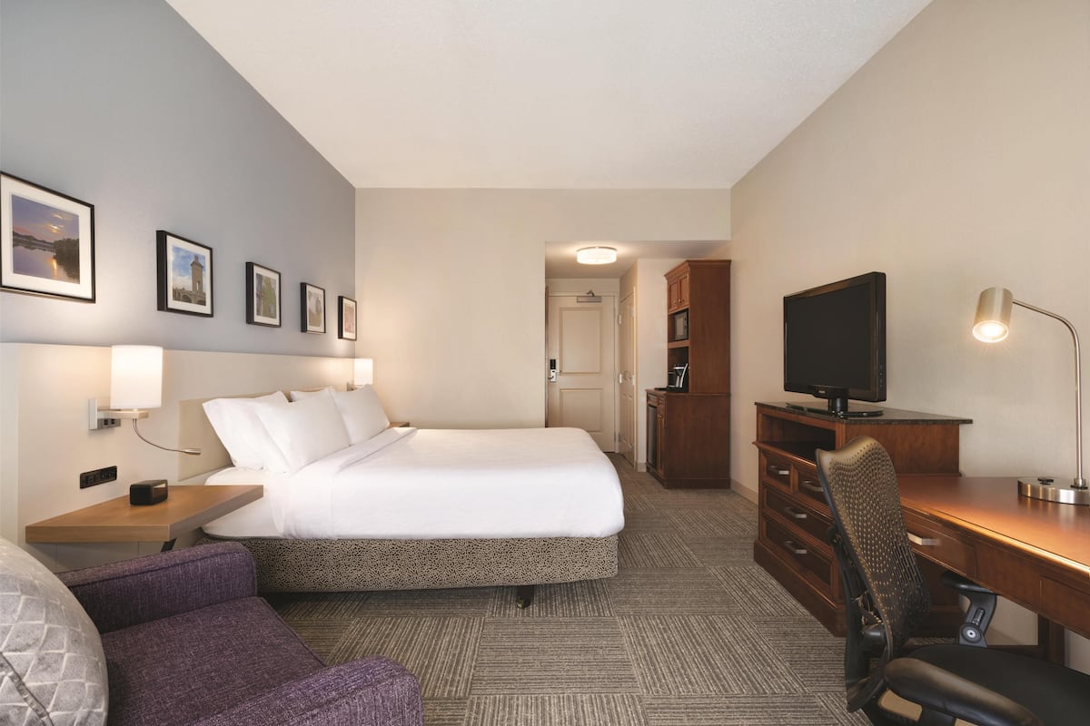 Quarto de hotel com cama, escrivaninha, cadeira, televisão e quadros na parede.