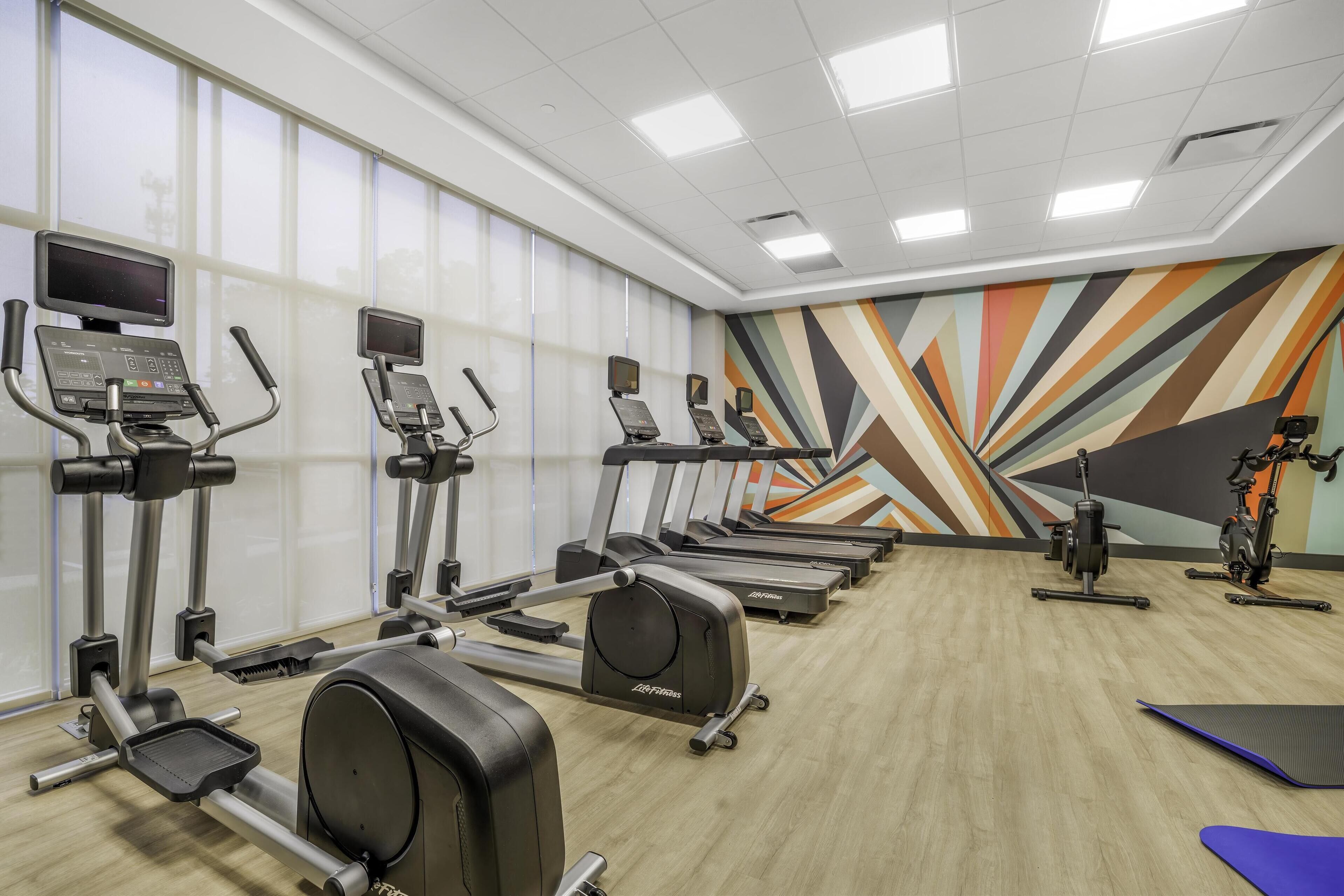 Une salle de sport équipée de tapis de course, de vélos elliptiques et ornée d’une fresque murale aux couleurs vives.